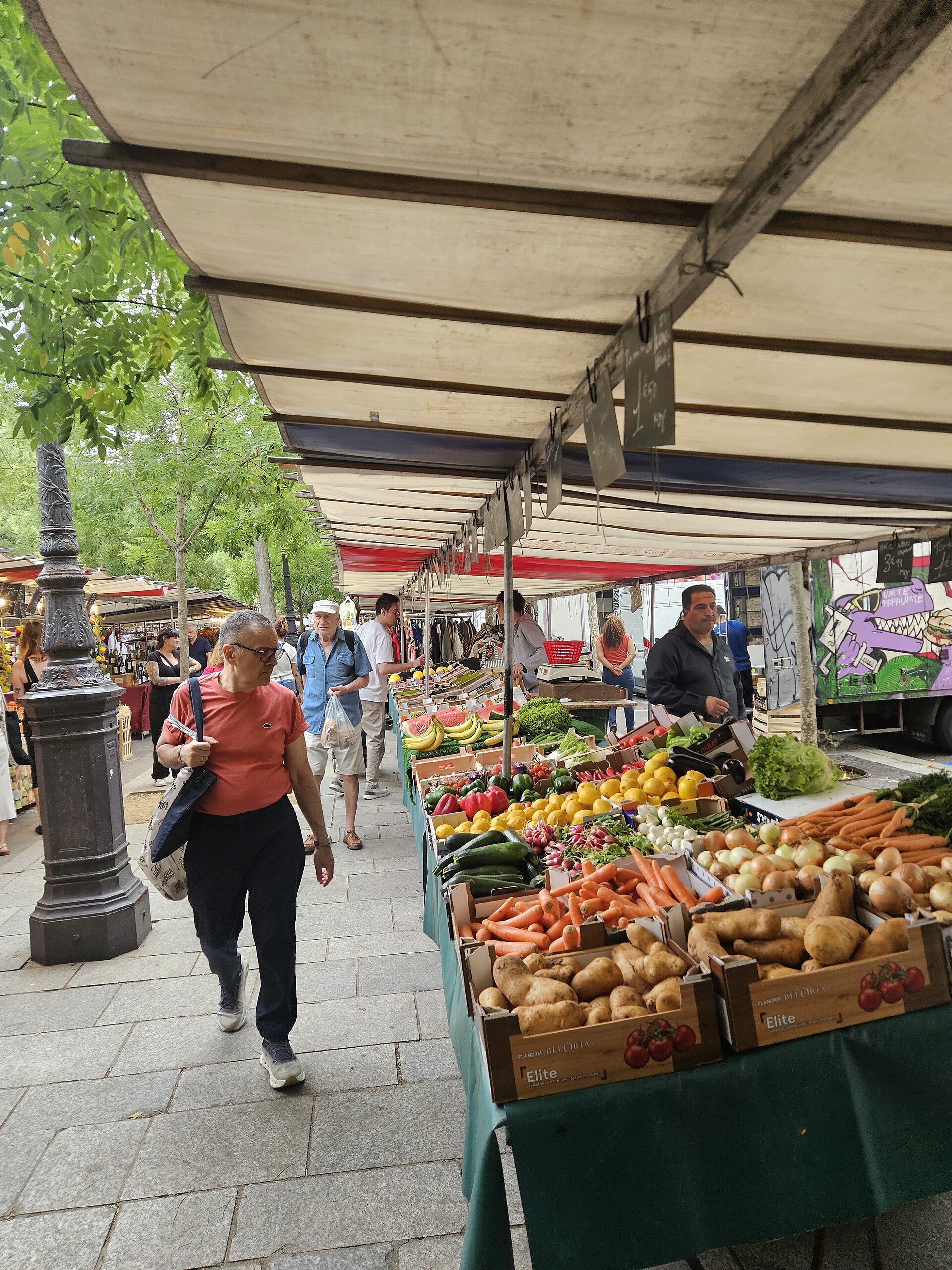 Awnings and produce stalls of the Bastille market on Boulevard Richard-Lenoir in Paris, with shoppers walking past crates of vegetables