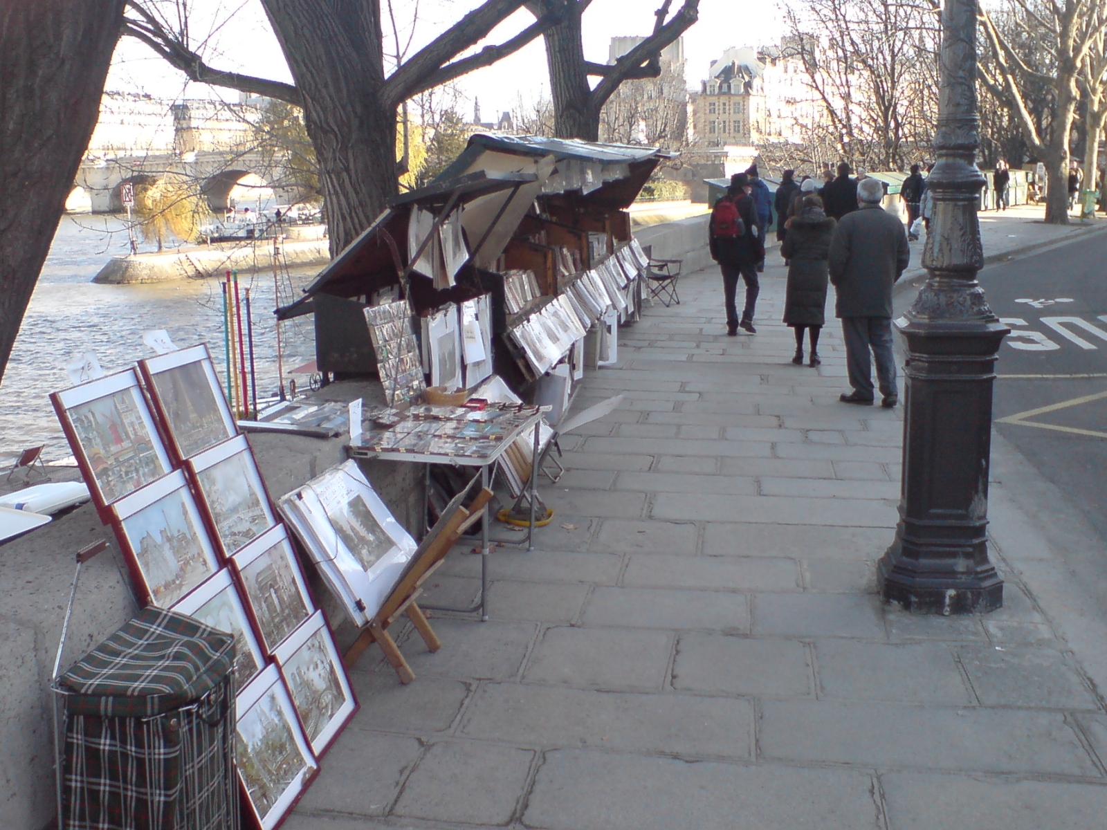 Green bouquiniste boxes open along a Seine quai in Paris, with framed prints propped against the wall and pedestrians walking past