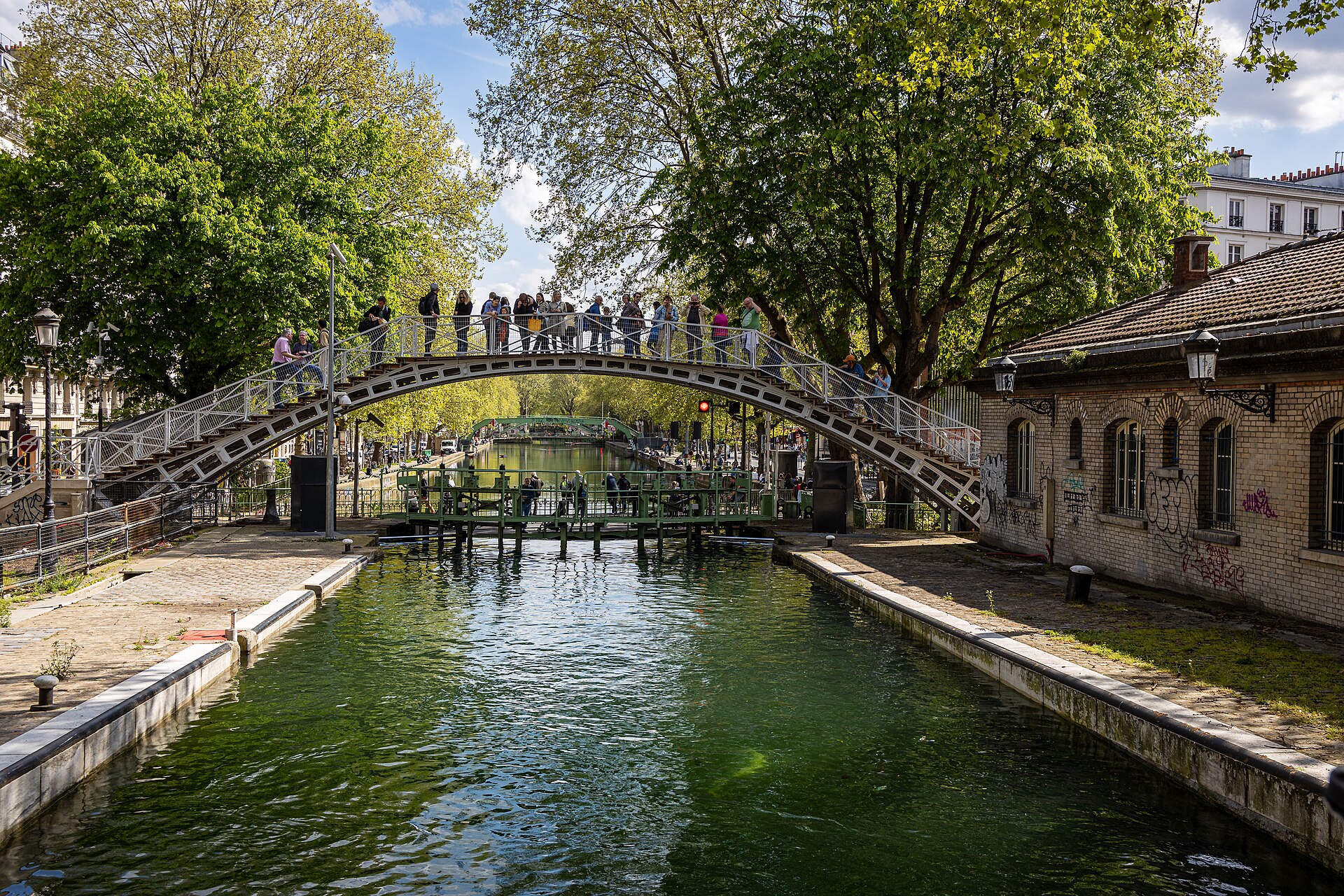 An arched iron footbridge over the Canal Saint-Martin in Paris, with plane trees, a lock and people standing on the bridge