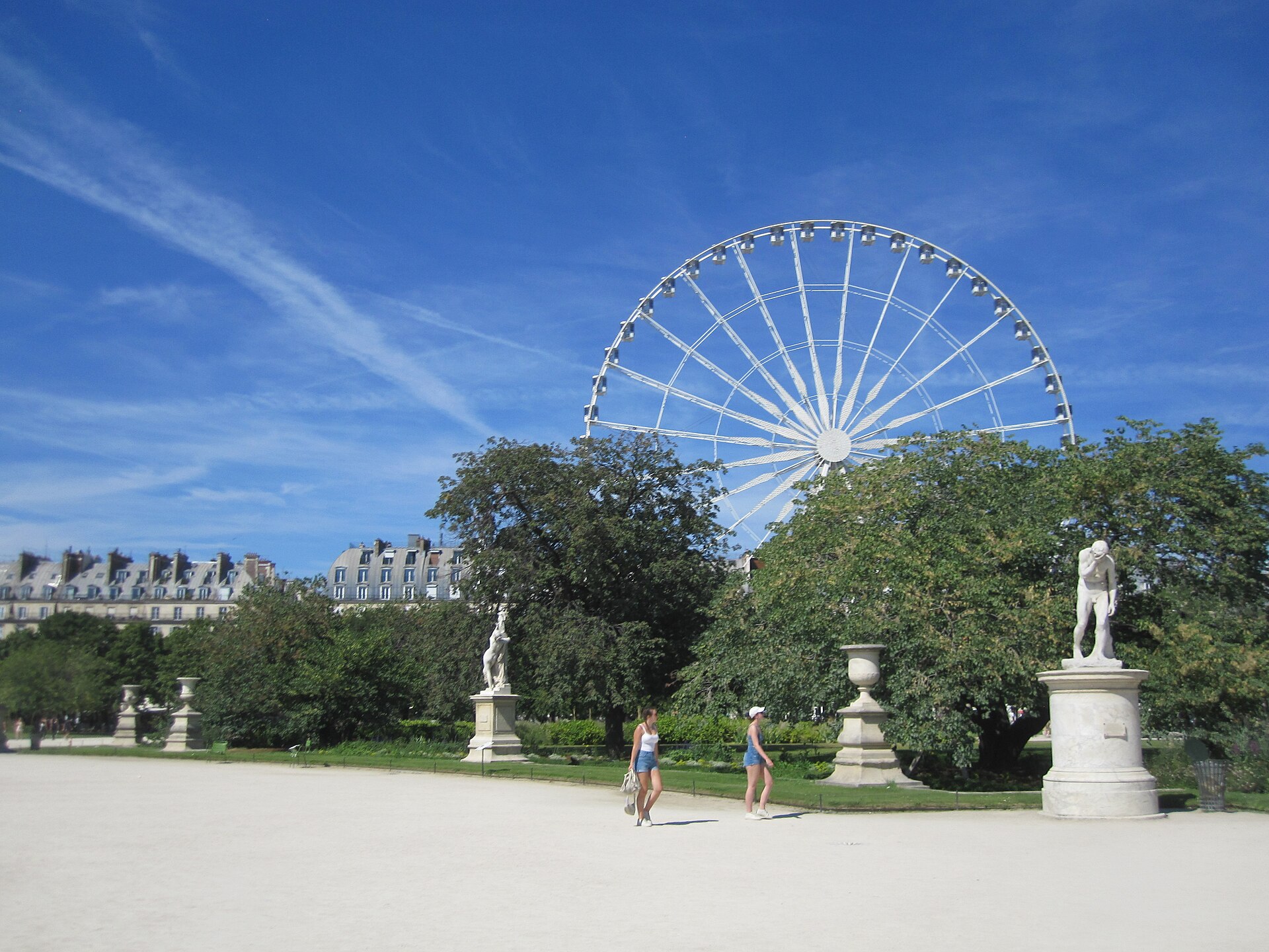 The white Ferris wheel of the Tuileries summer funfair rising behind trees and classical statues on a sunny day