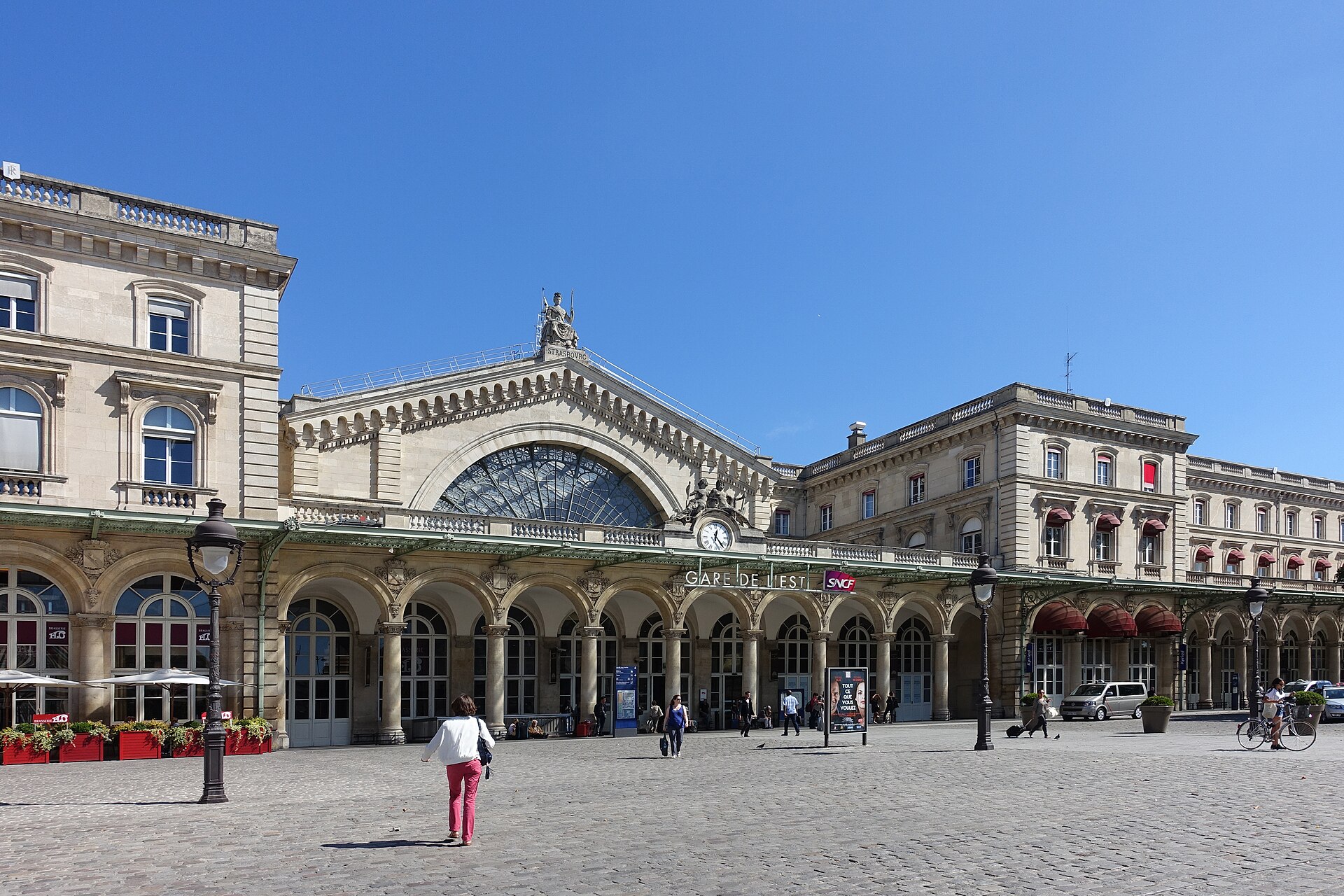 The long neoclassical façade of Gare de l'Est in Paris with its great arched central window, seen across a sunny cobbled forecourt