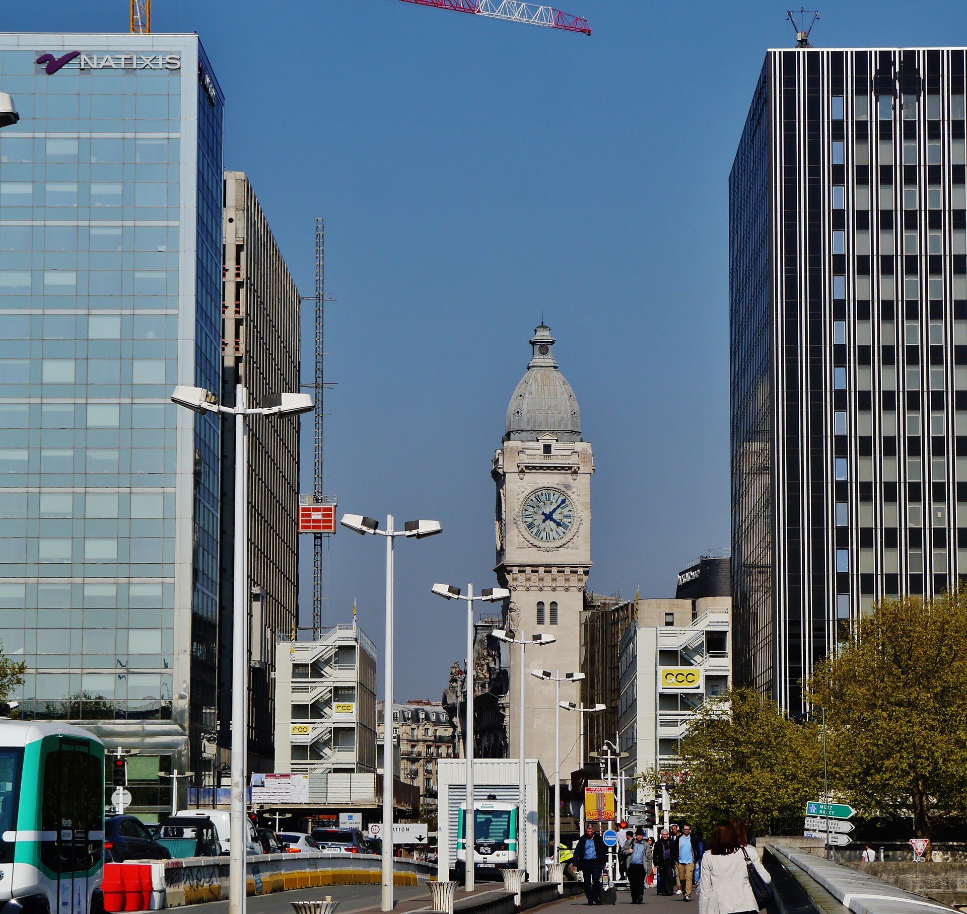 The clock tower of Gare de Lyon rising between modern office buildings under a blue sky, with a bus and pedestrians in front