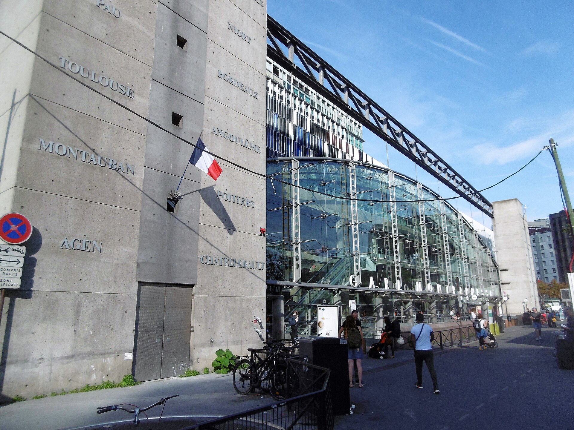 The modern glass entrance of Gare Montparnasse beside a concrete wall carved with the names of destinations including Toulouse, Montauban and Agen