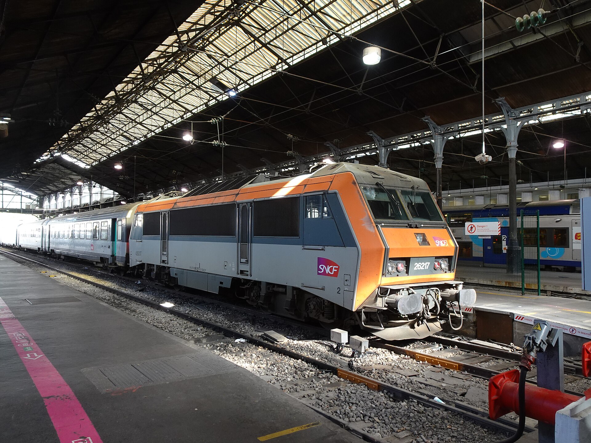 An orange and grey SNCF electric locomotive standing at a platform under the iron and glass roof of Gare Saint-Lazare