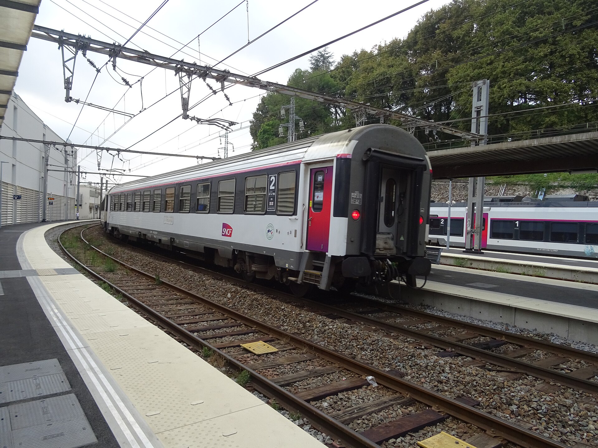 A white and grey Intercités de Nuit sleeper carriage with red doors standing at a curved station platform