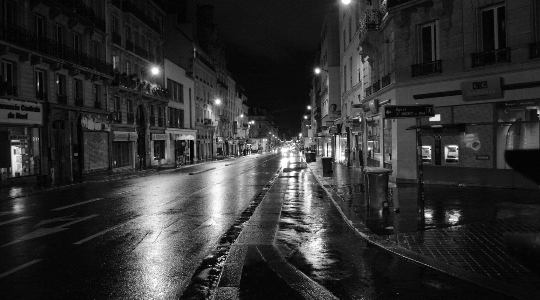 A wet Paris street at night in black and white, with lit street lamps and shuttered shopfronts
