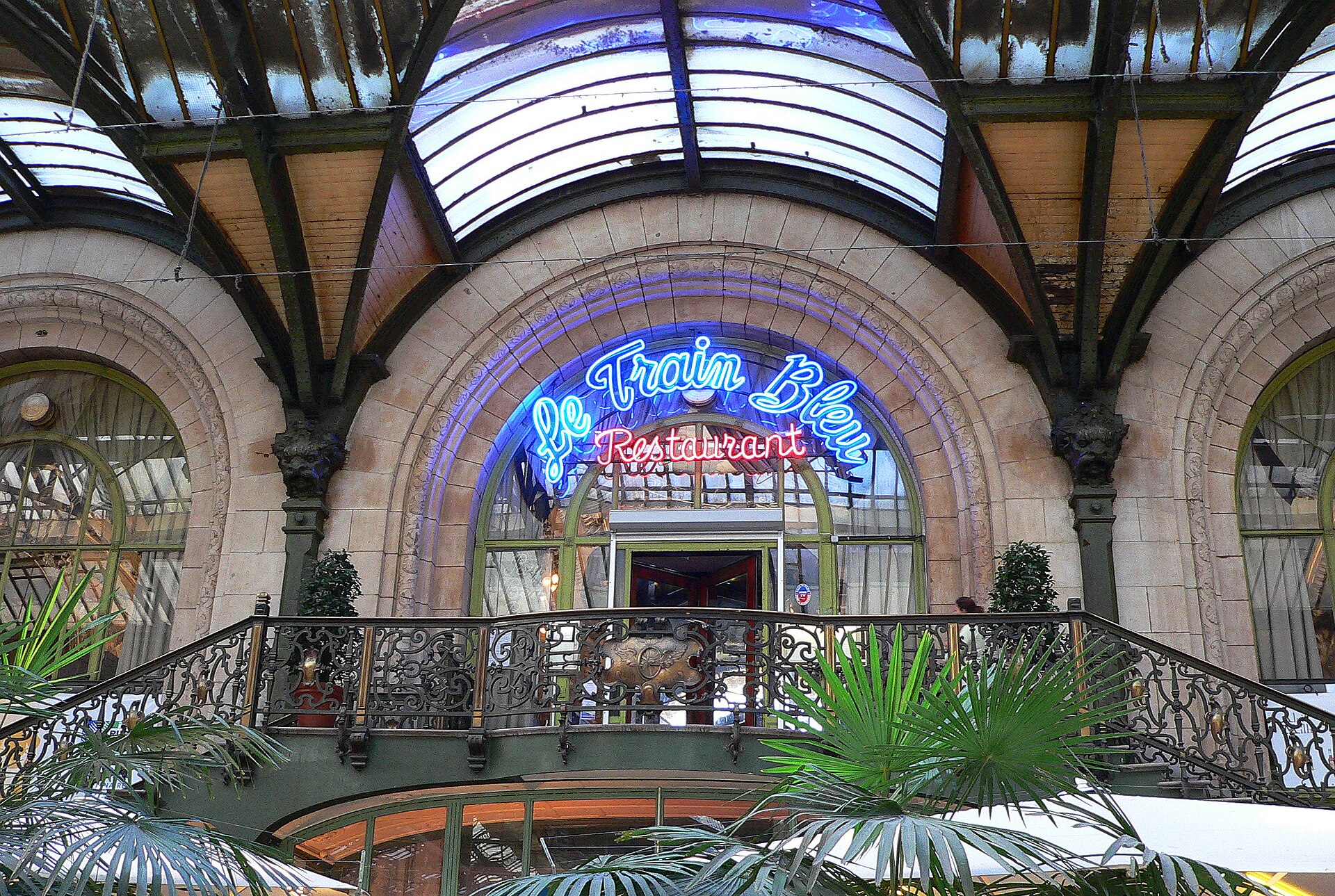 The ornate entrance of Le Train Bleu restaurant above the concourse at Gare de Lyon, with a blue neon sign, arched windows and wrought-iron railings