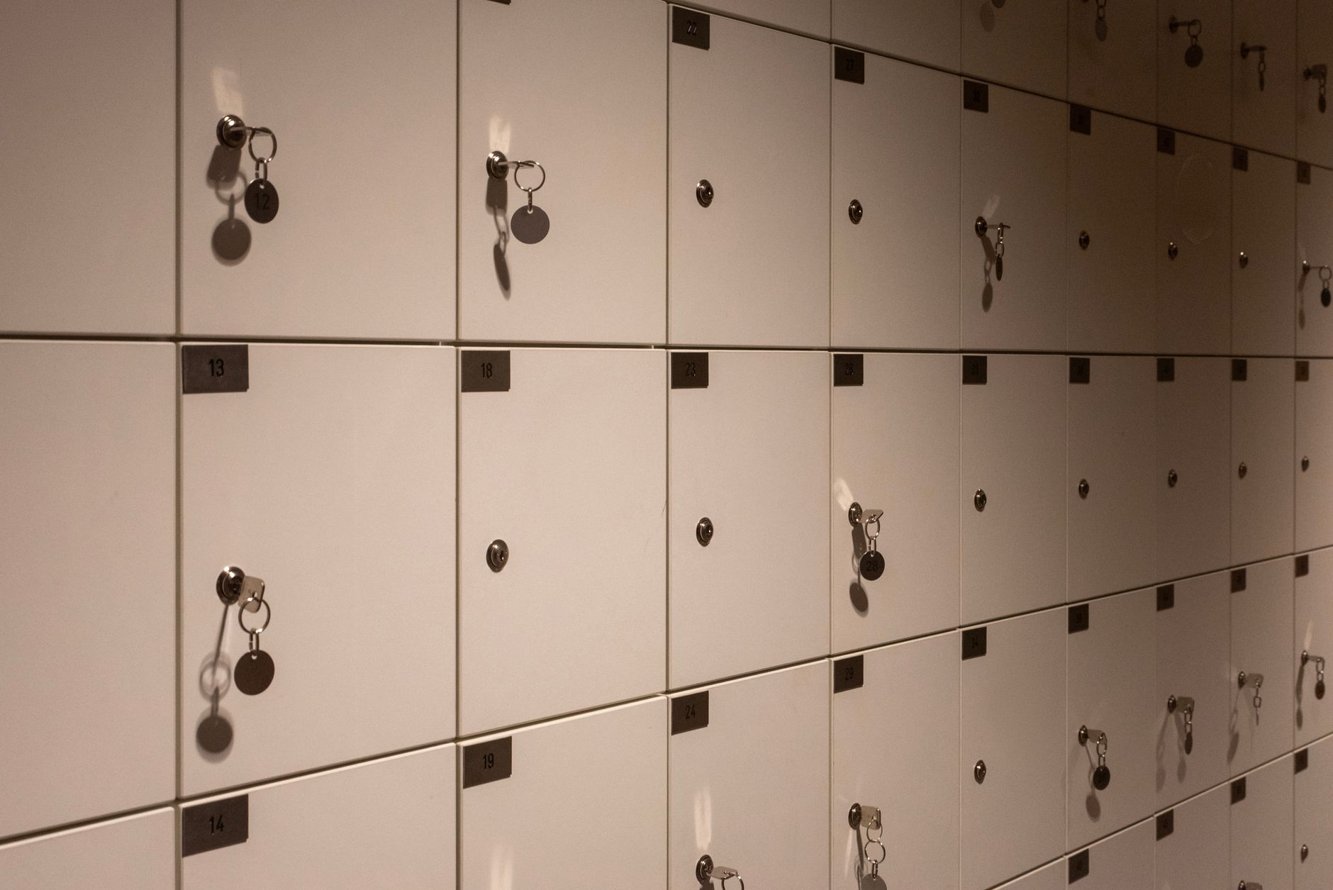 A long wall of pale grey metal lockers with numbered doors, several with keys and round key tags hanging from the locks