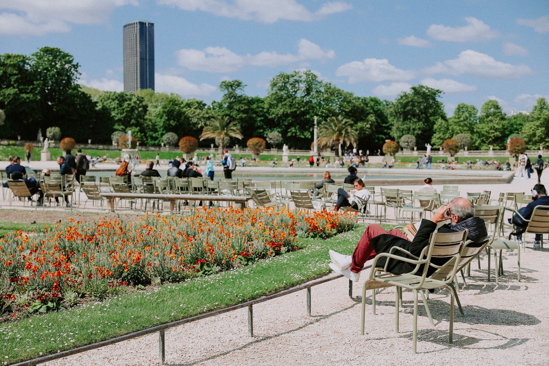 Visitors lounging on green metal chairs beside bright orange and red flowerbeds in the Jardin du Luxembourg, with the Montparnasse tower visible above the trees