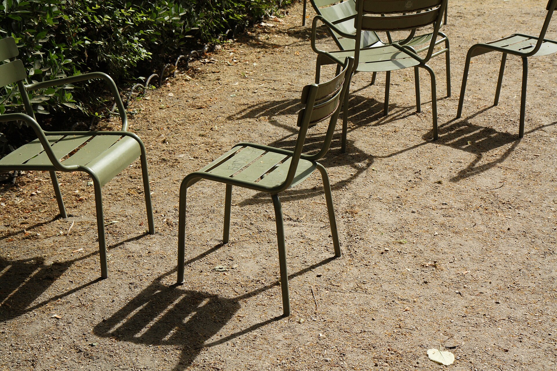 A cluster of empty sage-green metal chairs casting long shadows on the gravel in the Jardin du Luxembourg