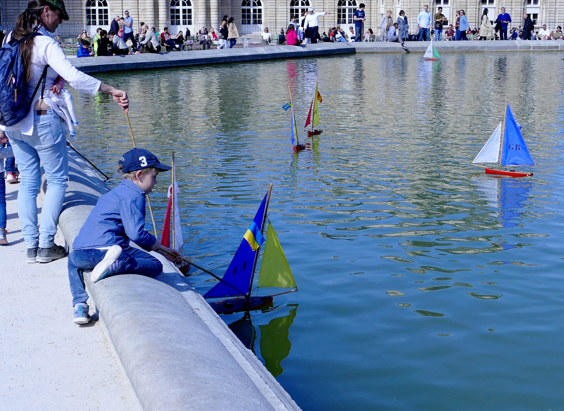 A small boy in a cap leaning over the stone edge of the Grand Bassin to push a toy sailboat with a stick, with other colourful boats on the water