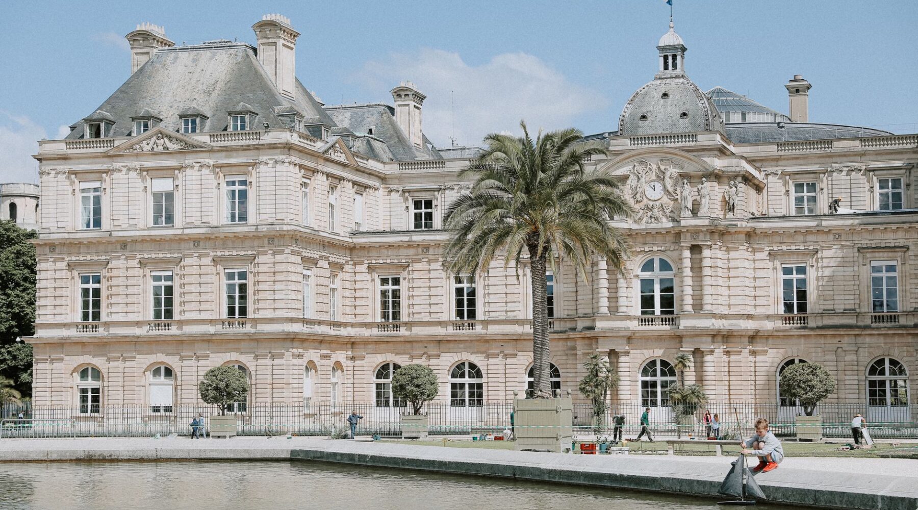 The Luxembourg Palace with its central dome and French flag seen across a pond, with a palm tree in a planter on the terrace