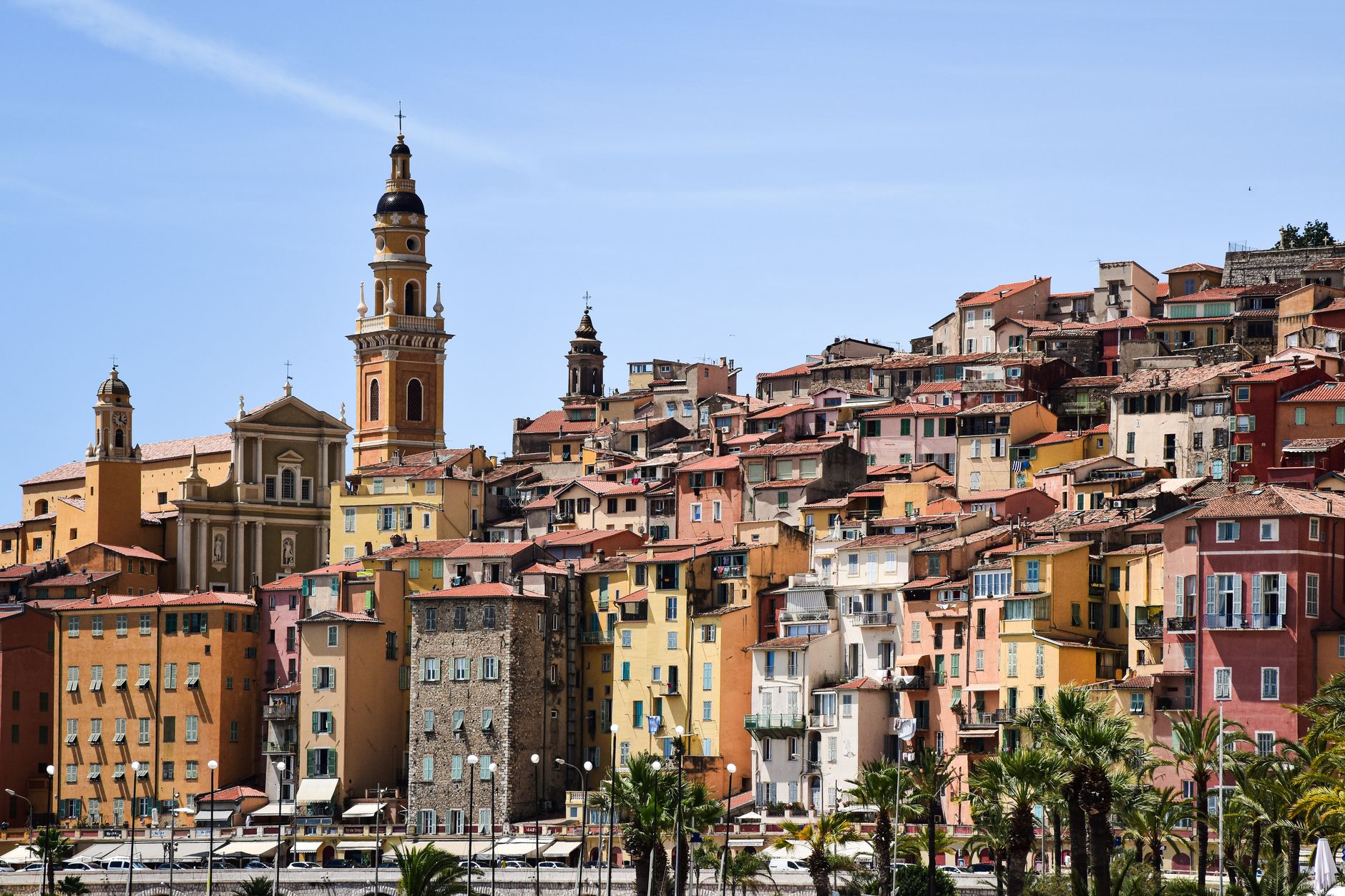 The pastel-coloured houses of Menton's old town rising up a hillside to a baroque church bell tower, with palm trees along the waterfront