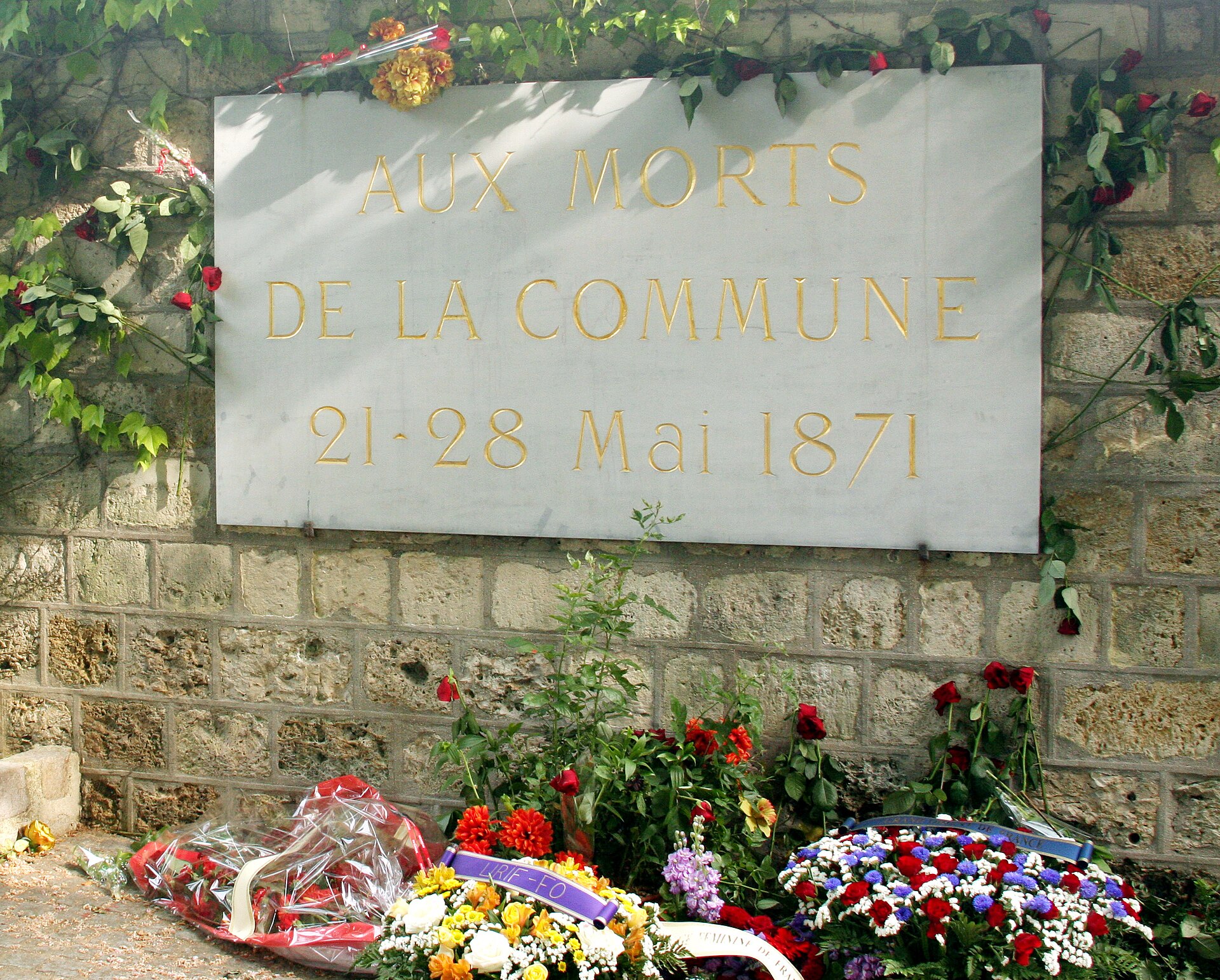 The stone plaque of the Mur des Fédérés reading Aux Morts de la Commune 21-28 Mai 1871, set in a rough stone wall with wreaths and flowers below