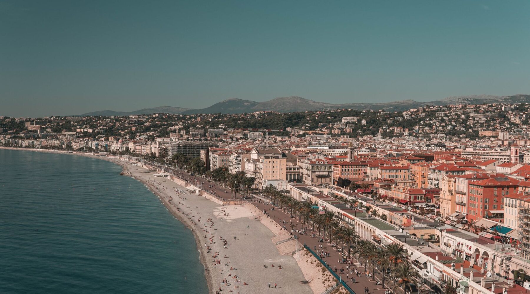 Aerial view of the curving pebble beach and turquoise water of the Baie des Anges in Nice, with the city and hills behind
