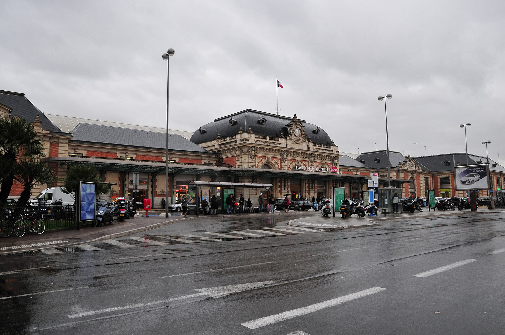 The ornate 19th-century façade of Nice-Ville railway station with its central clock pavilion, seen across a wet street on a grey day