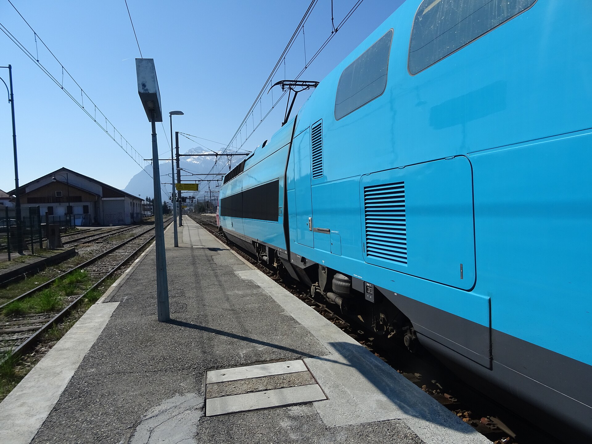The bright blue double-decker carriage of a OUIGO low-cost high-speed train standing at a station platform under a clear sky