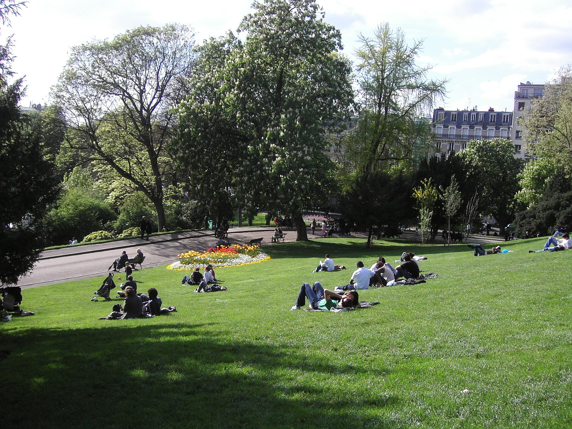 People lying and sitting on a sunny sloping lawn in the Parc des Buttes-Chaumont, with tall trees and a flowerbed behind them