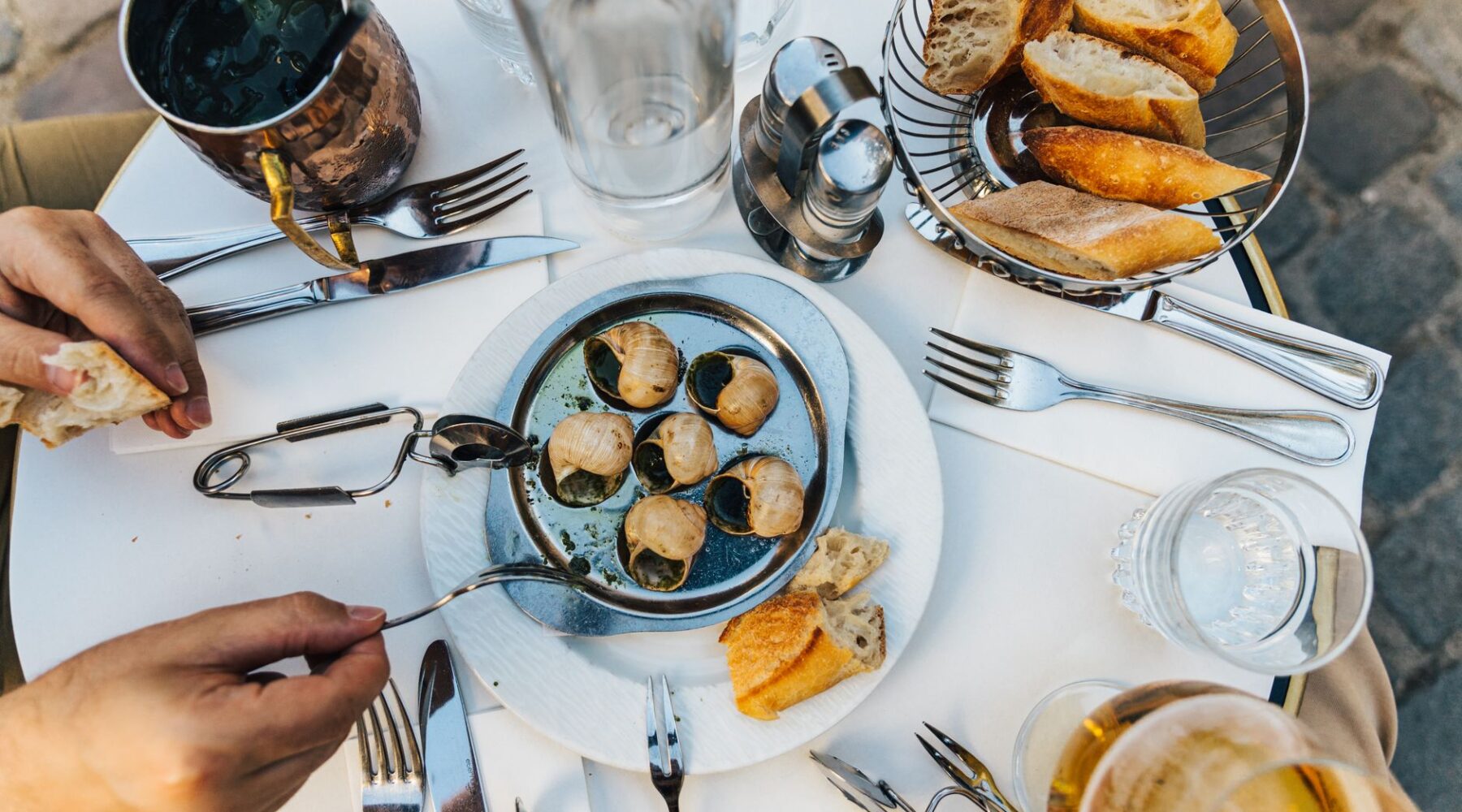An overhead view of a marble bistro table with a plate of escargots, tongs, a basket of baguette and glasses of wine