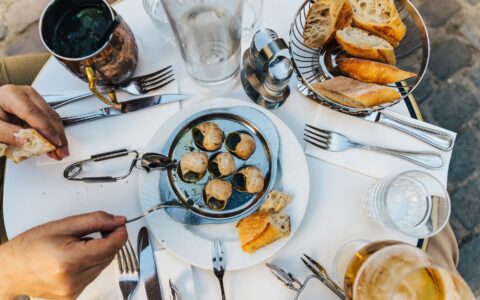An overhead view of a marble bistro table with a plate of escargots, tongs, a basket of baguette and glasses of wine