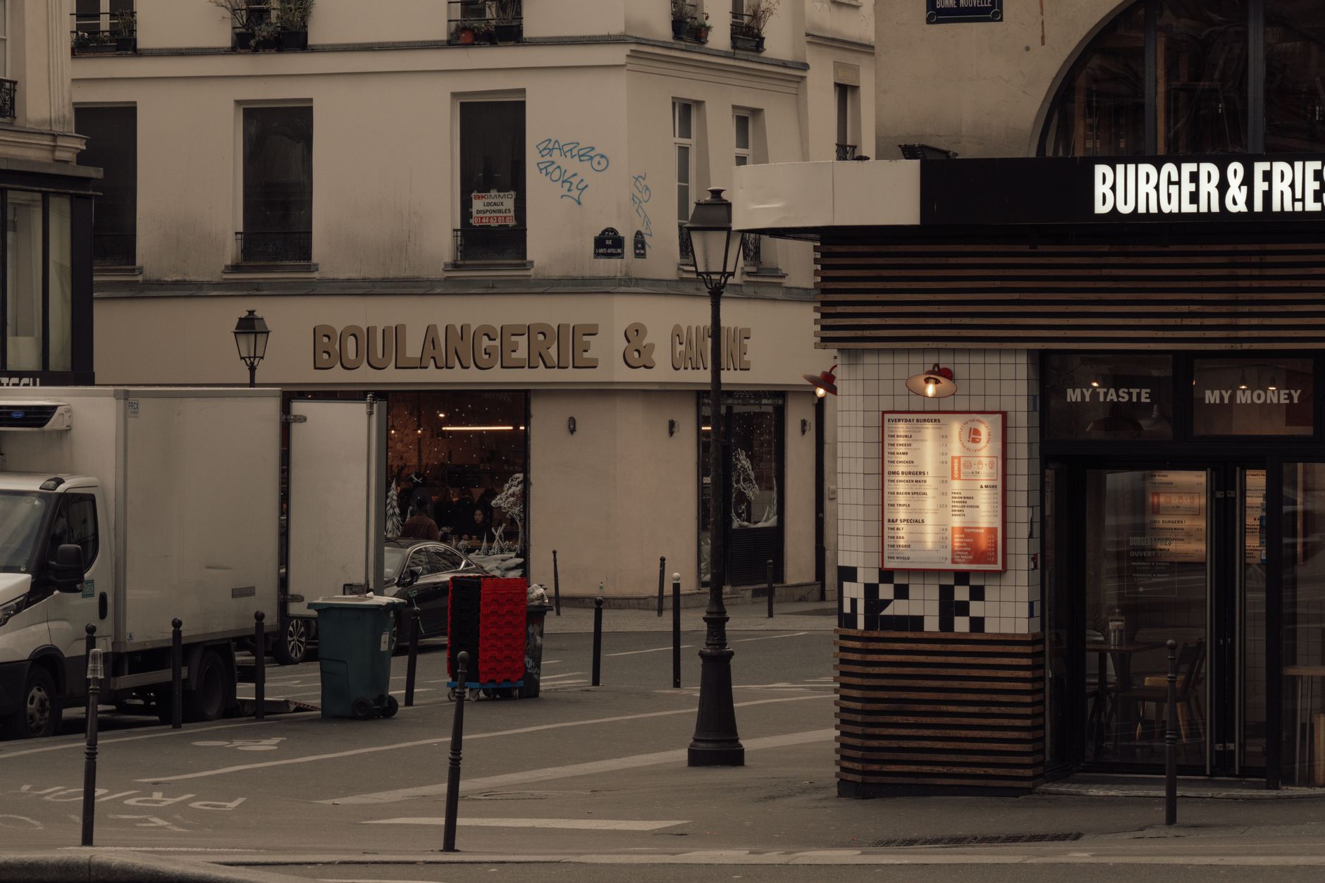 A Paris street corner at dusk with a boulangerie sign next to a modern burger restaurant, a delivery lorry parked outside