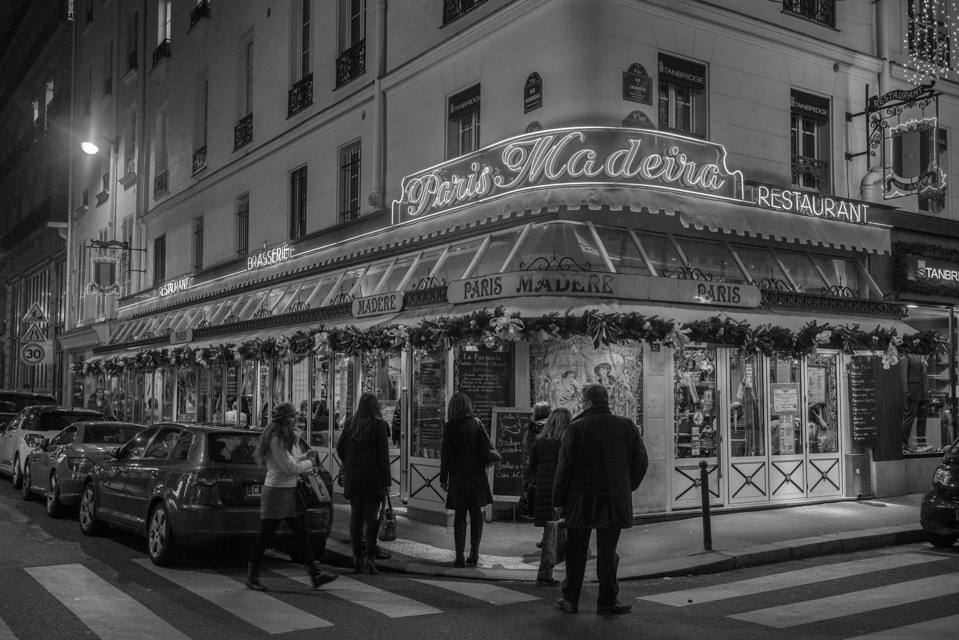 People reading the chalked formule menu outside a lit Paris brasserie on a street corner at night