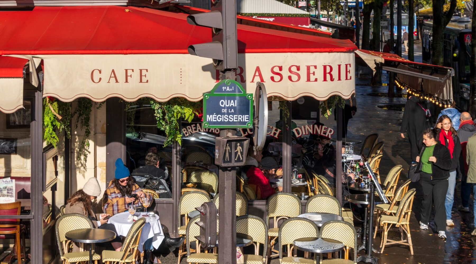 A Paris café-brasserie on a street corner with a red awning, rattan chairs and people sitting out on an autumn afternoon