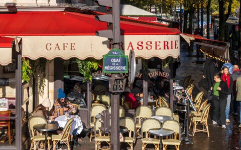 A Paris café-brasserie on a street corner with a red awning, rattan chairs and people sitting out on an autumn afternoon