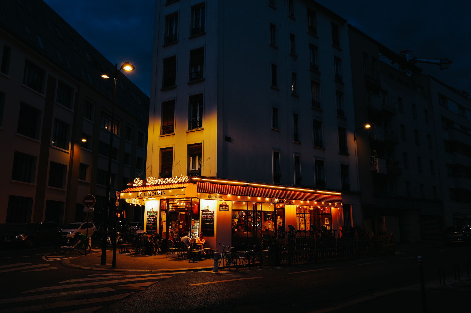 A brightly lit corner brasserie with an orange awning and outdoor tables on a dark residential Paris street at night
