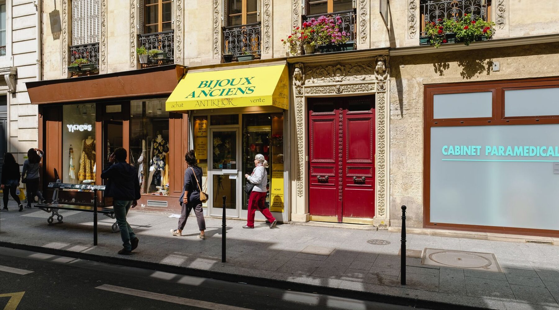 A Paris street with a small antique jewellery shop under a yellow awning, a boutique and people walking past