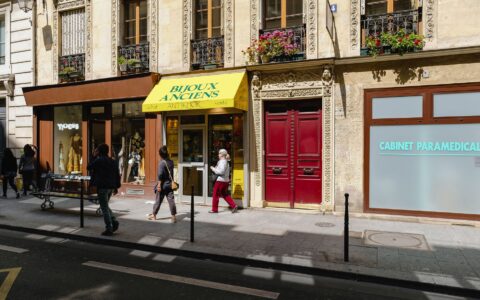 A Paris street with a small antique jewellery shop under a yellow awning, a boutique and people walking past