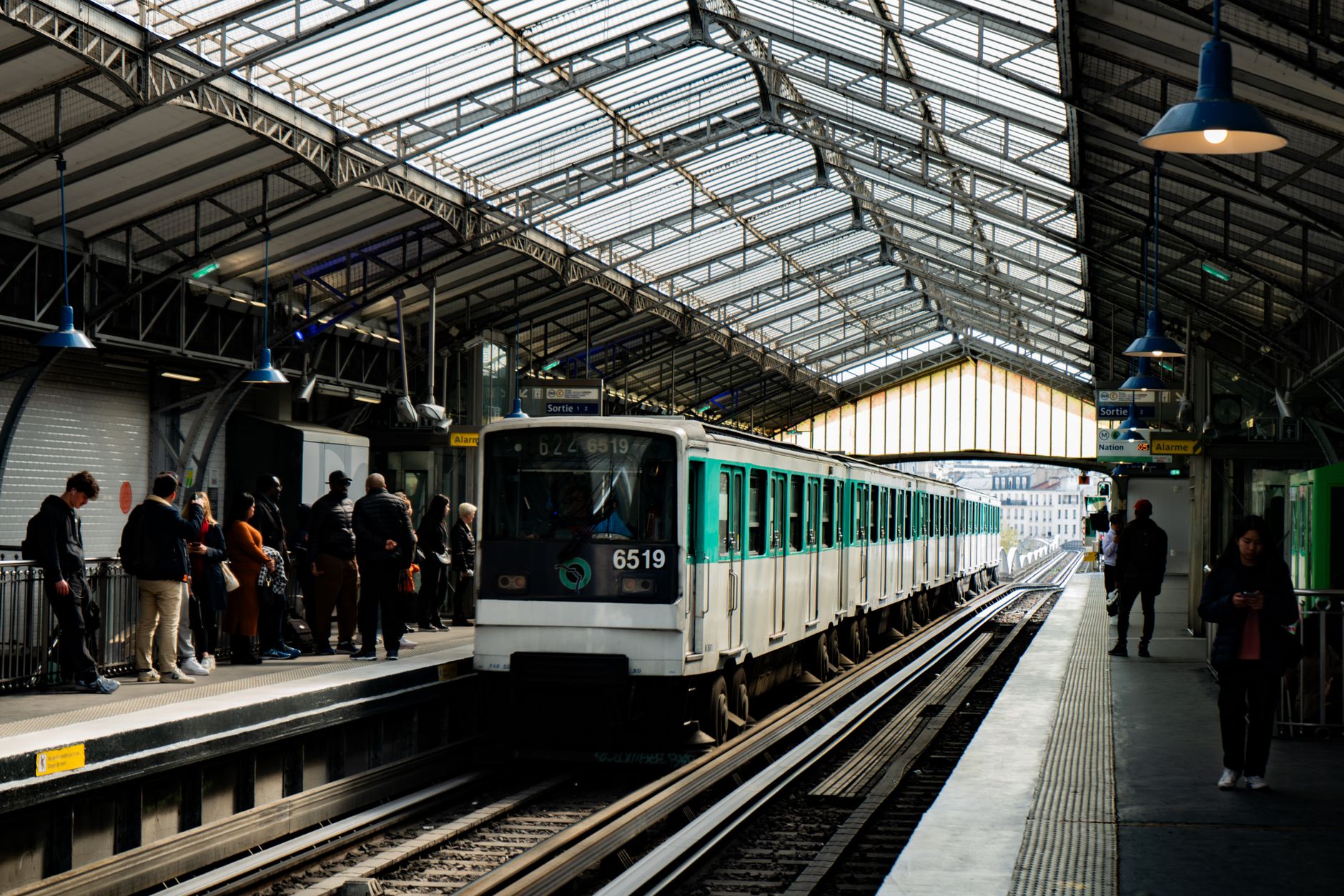 Passengers waiting close to the platform edge as a green and white Paris Metro train arrives at a glass-roofed elevated station