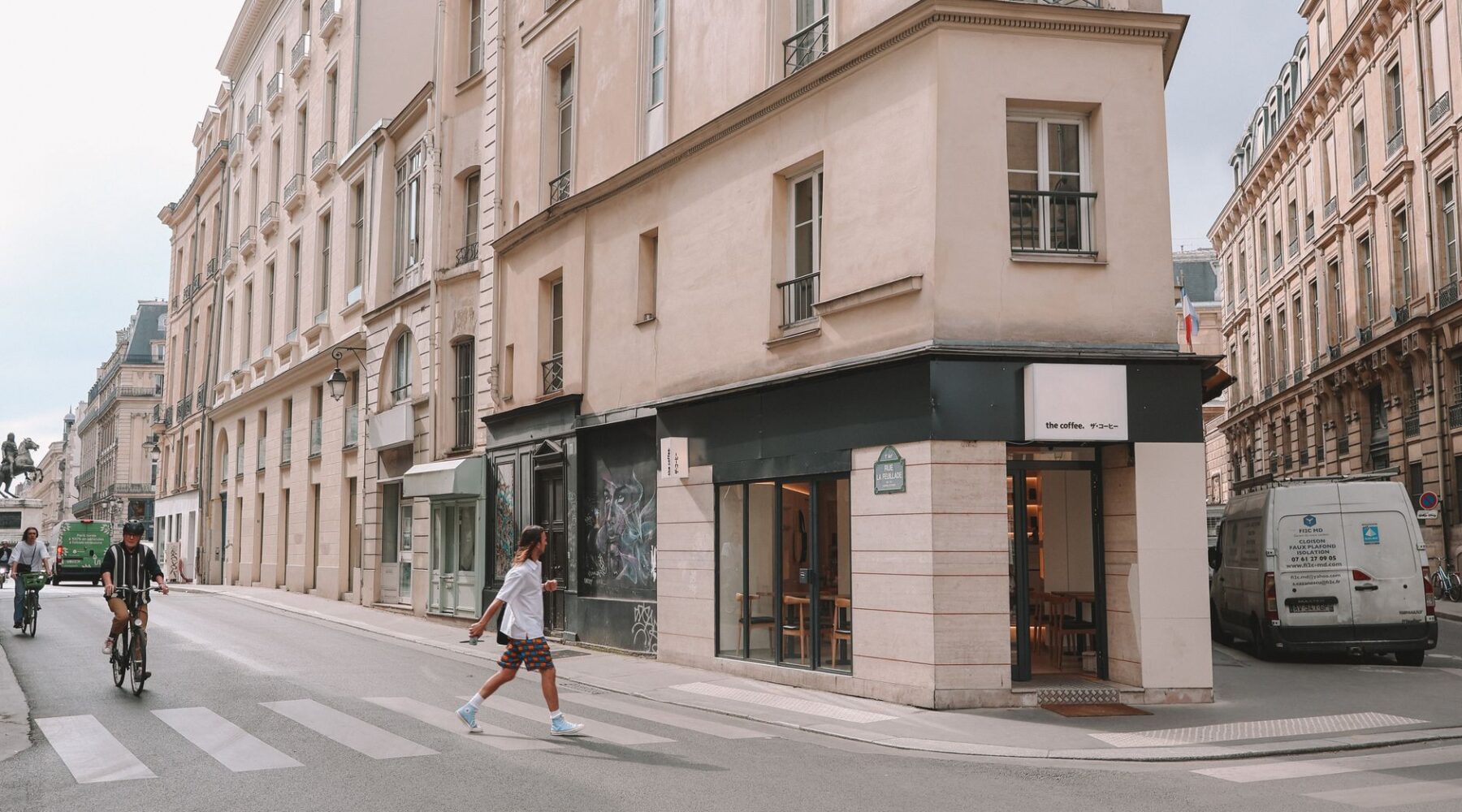 An ordinary Paris street corner with a small coffee shop, a cyclist and a delivery van outside cream stone buildings