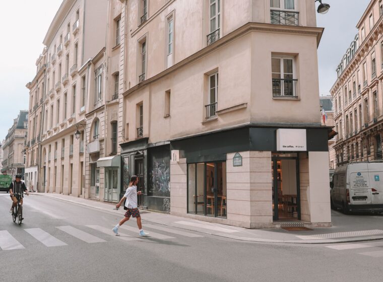 An ordinary Paris street corner with a small coffee shop, a cyclist and a delivery van outside cream stone buildings