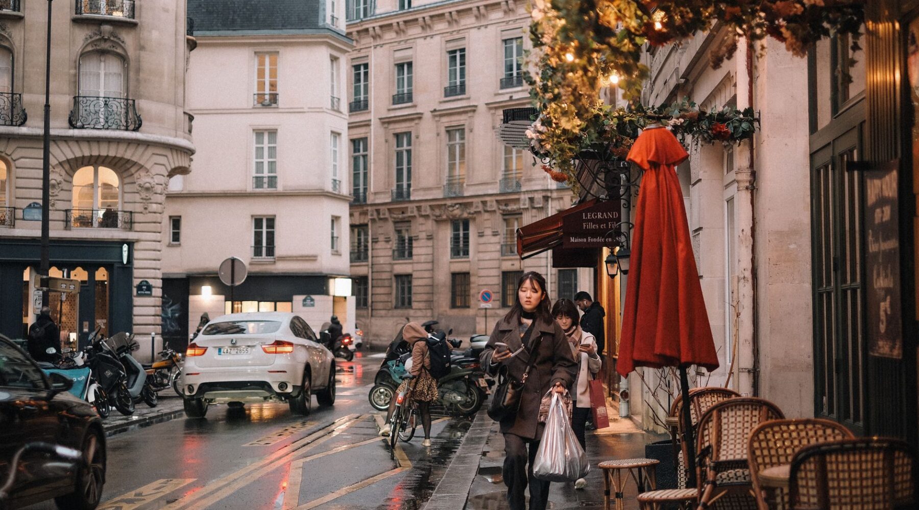 Two people walking along a wet Paris street looking at their phones, past a cafe terrace with a red parasol