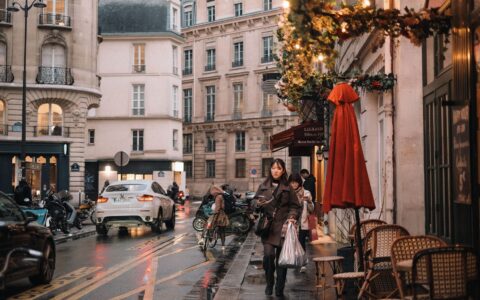 Two people walking along a wet Paris street looking at their phones, past a cafe terrace with a red parasol