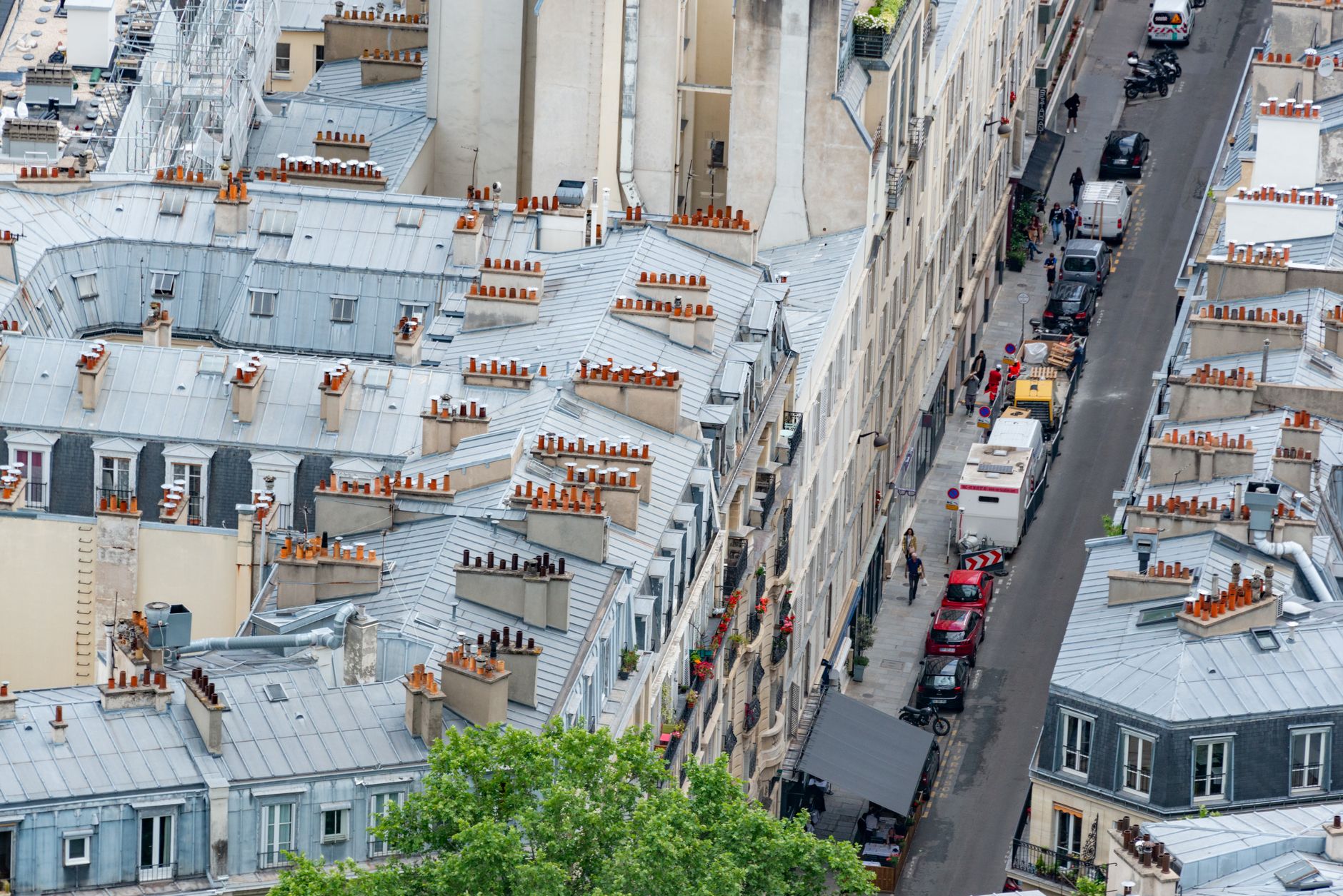High-angle view over Parisian zinc rooftops and chimney pots down onto an ordinary narrow street with delivery vans and pedestrians