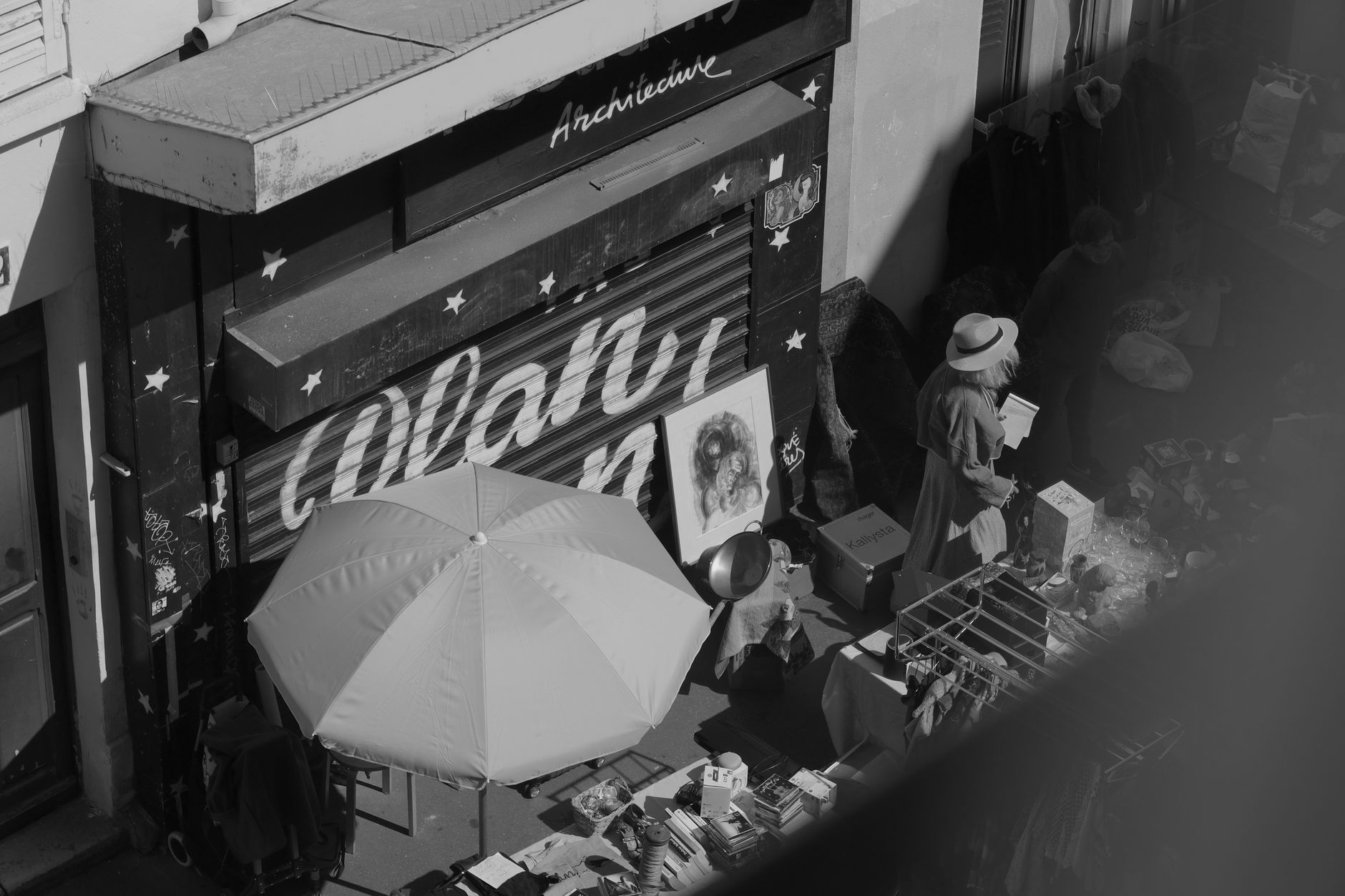An overhead view of a Paris street brocante with a parasol, framed pictures and a woman browsing a table of bric-a-brac