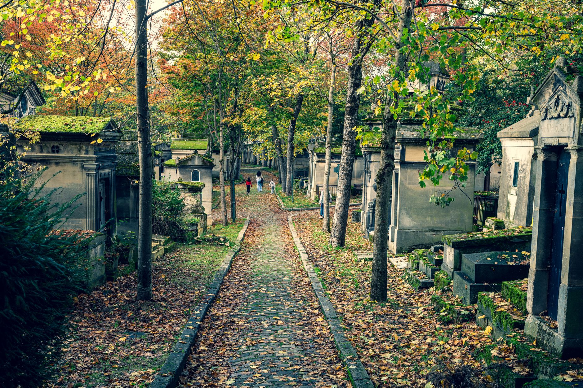 Tombs and family chapels along a narrow cobbled path at Père Lachaise, with autumn leaves on the ground and grey sky above