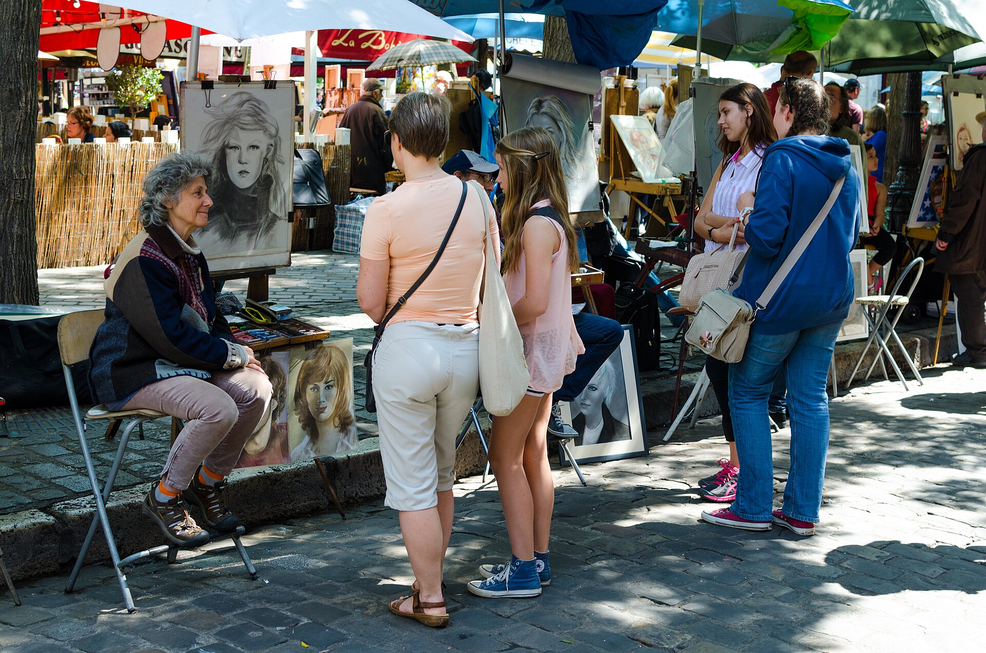 Portrait artists at easels on Place du Tertre in Montmartre, with visitors standing and watching a charcoal drawing being made