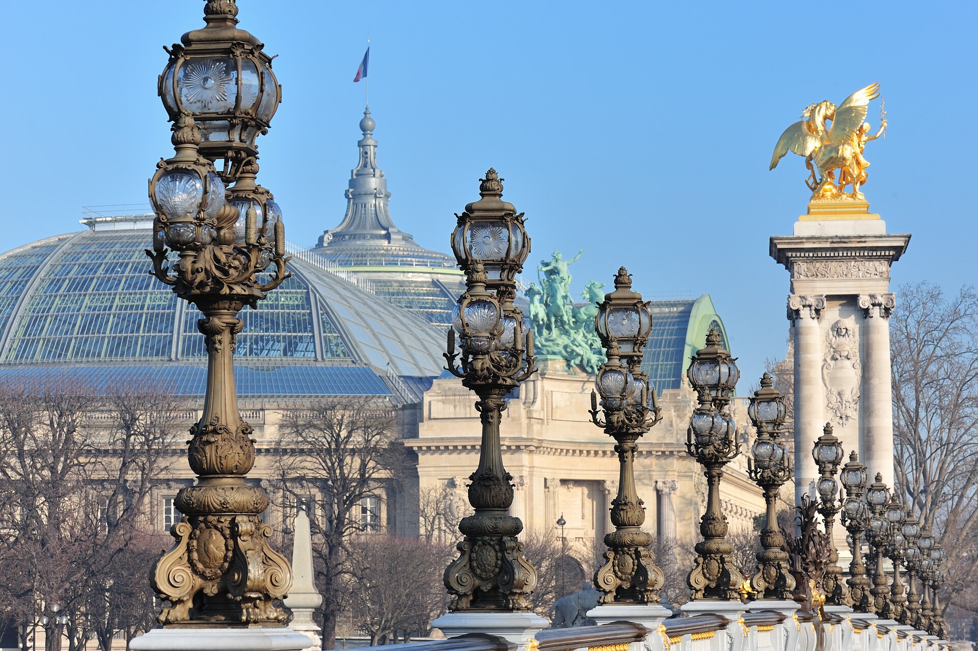 The ornate lamp standards and gilded winged statue of the Pont Alexandre III in Paris, with the glass dome of the Grand Palais behind