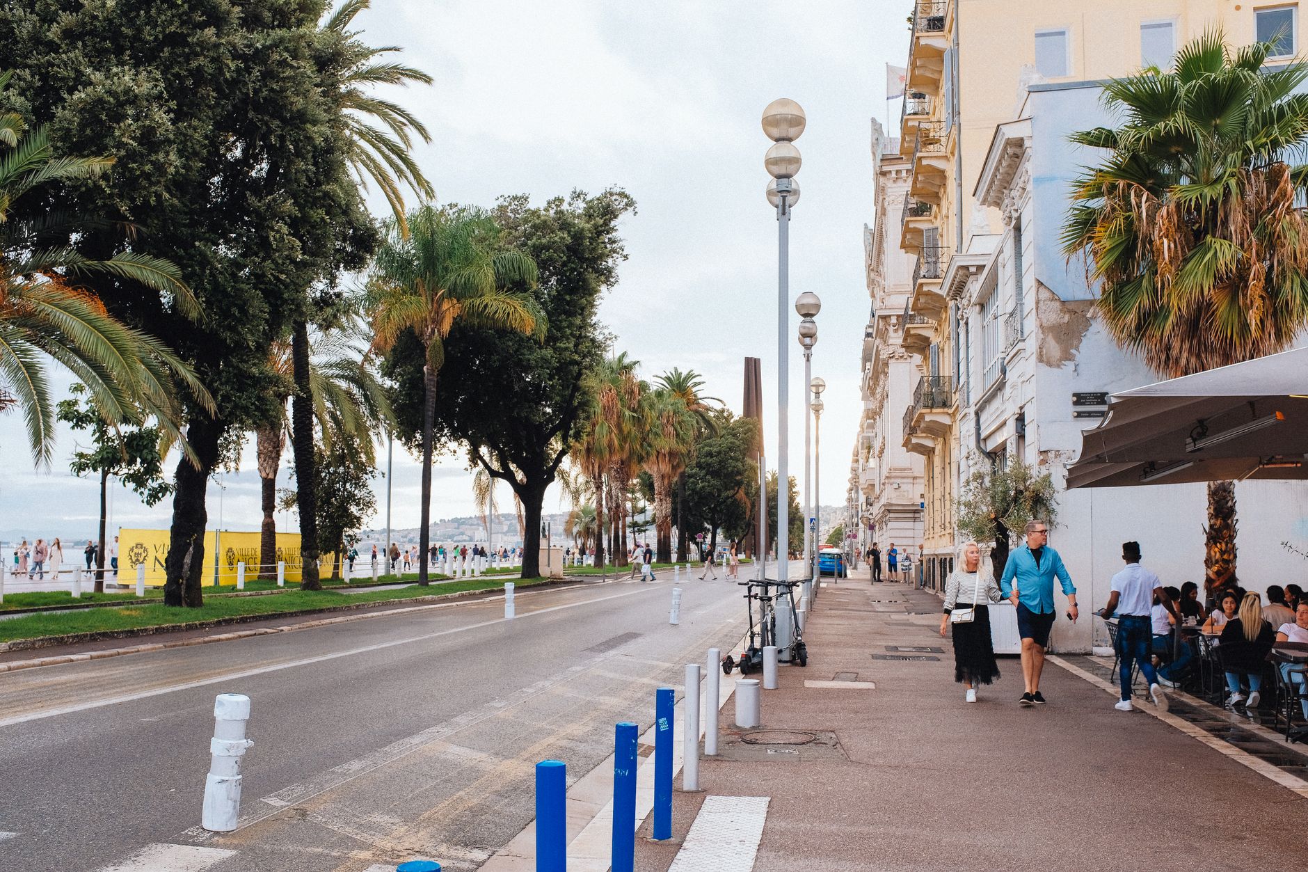 People walking along the palm-lined Promenade des Anglais in Nice on a sunny day, beside the seafront road and café terraces