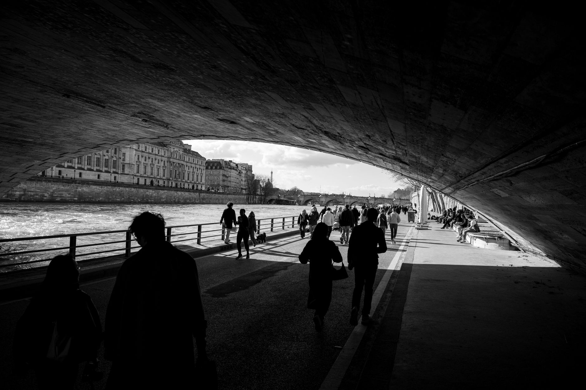 People walking along the lower Seine quay beneath a stone bridge arch in Paris, with the Pont Neuf and the Eiffel Tower visible beyond