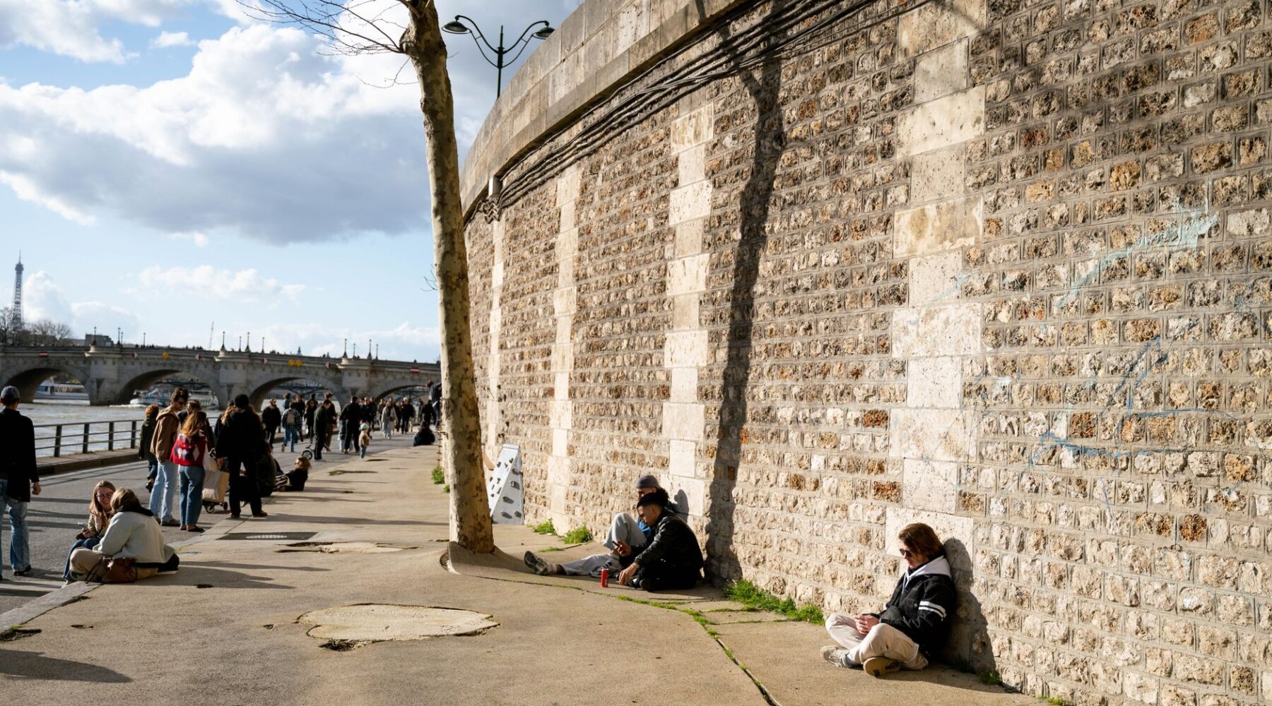 People sitting alone in the sun along the stone wall of the lower Seine quay in Paris, with the Pont Neuf and the Eiffel Tower behind