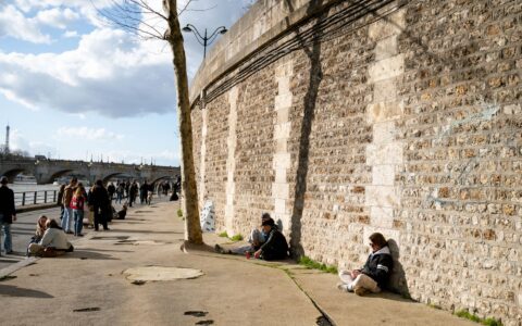 People sitting alone in the sun along the stone wall of the lower Seine quay in Paris, with the Pont Neuf and the Eiffel Tower behind