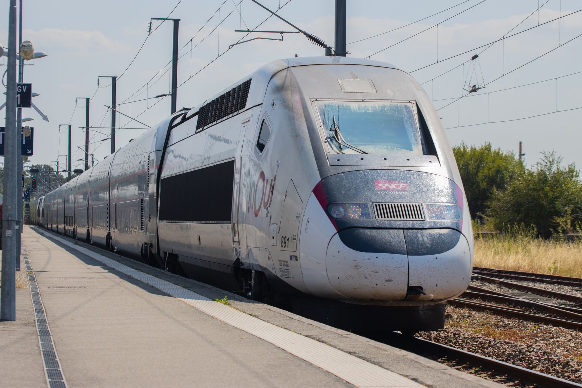 A white and grey TGV INOUI high-speed train arriving at a station platform in the French countryside on a sunny day