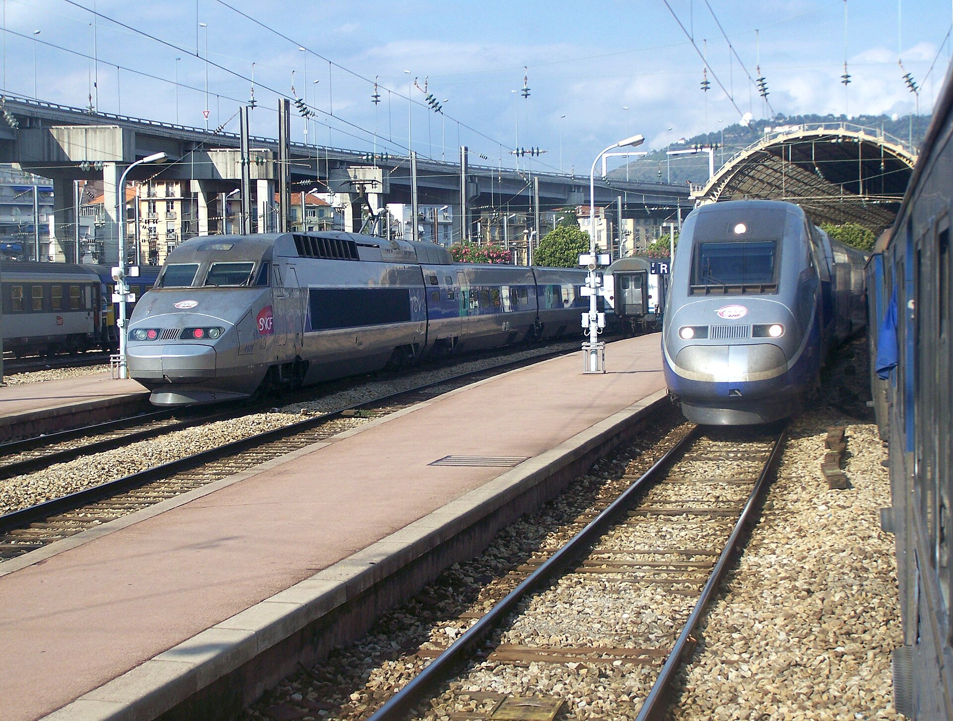 Two silver and blue TGV high-speed trains standing at the platforms of Nice-Ville station under a sunny sky, with hills behind
