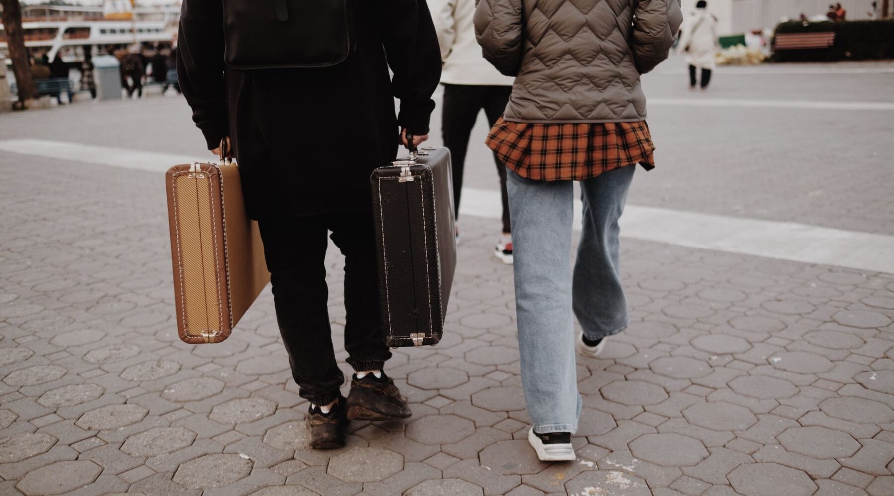Two travellers seen from behind walking across a paved square, each carrying a vintage hard-shell suitcase