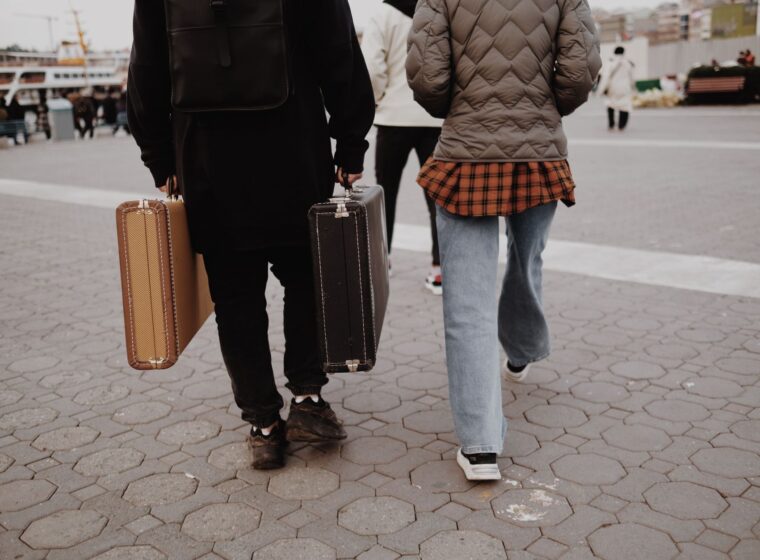 Two travellers seen from behind walking across a paved square, each carrying a vintage hard-shell suitcase