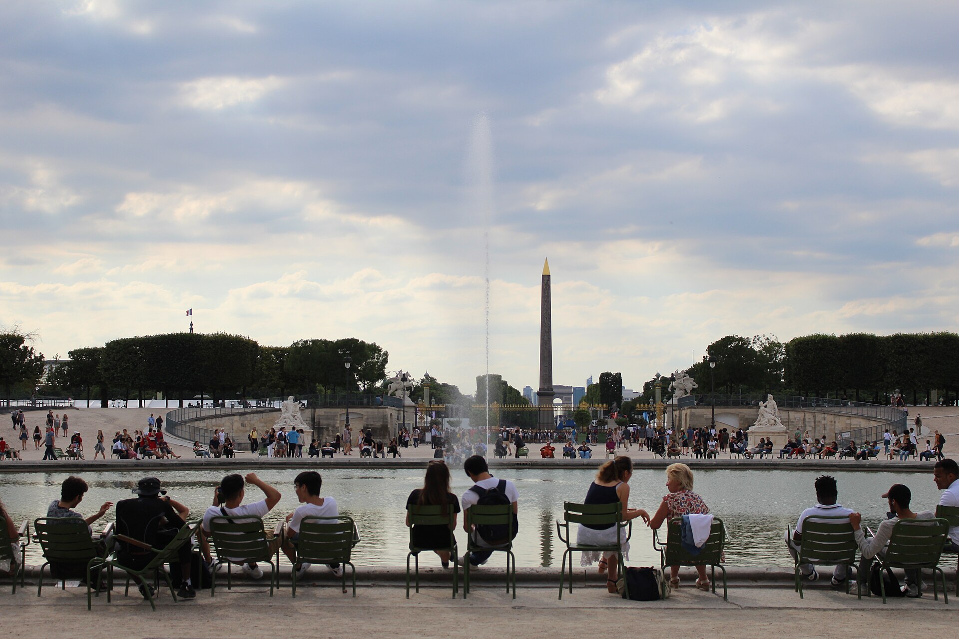 Visitors sitting on green metal chairs along the rim of the octagonal pond in the Tuileries, with a fountain jet and the Concorde obelisk in the distance
